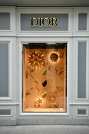 A dior storefront window display with illuminated golden floral art gold cutouts and hanging fine jewelry handbags