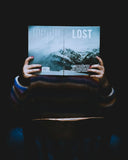 A person holds an open lost magazine with a snowy mountain cover featuring bold white lettering on gold jewelry.