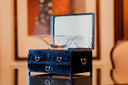 A sleek black lacquered jewelry box with gold-toned ring handles holding a clear glass bowl for fine jewelry storage.