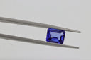 A deep blue, emerald-cut tanzanite gemstone held delicately between precision metal tweezers.