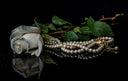 A strand of cream-colored pearls with an 18k gold clasp beside a wilted rose on a reflective surface in fine gold jewelry context
