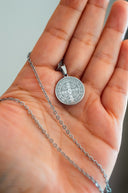 A hand holds a silver st. benedict medal necklace.