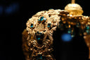 An ornate gold crown with square green gemstones against a dark background
