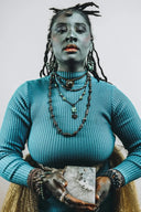 A woman wearing a teal ribbed turtleneck sweater and fine gold jewelry holds a rectangular white and gray art gold stone slab.