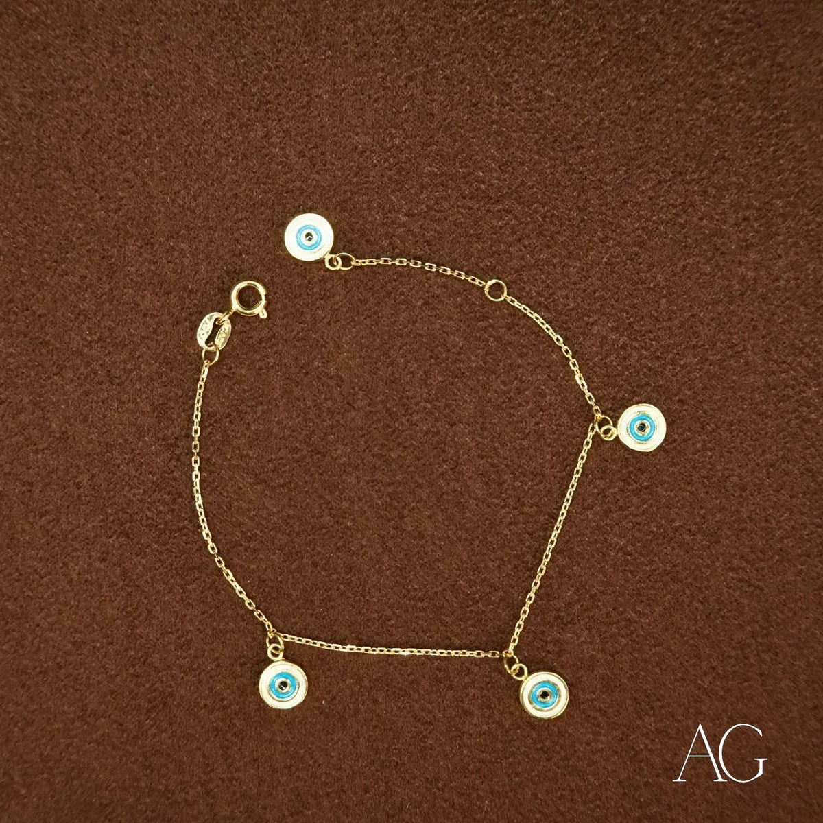 18k gold evil eye bracelet for kids with five dangling blue and white enamel charms, top-down view on brown surface