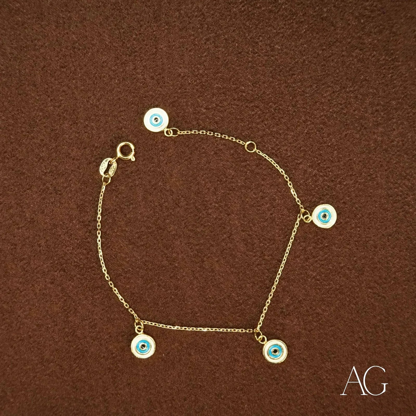 18k gold evil eye bracelet for kids with five dangling blue and white enamel charms, top-down view on brown surface