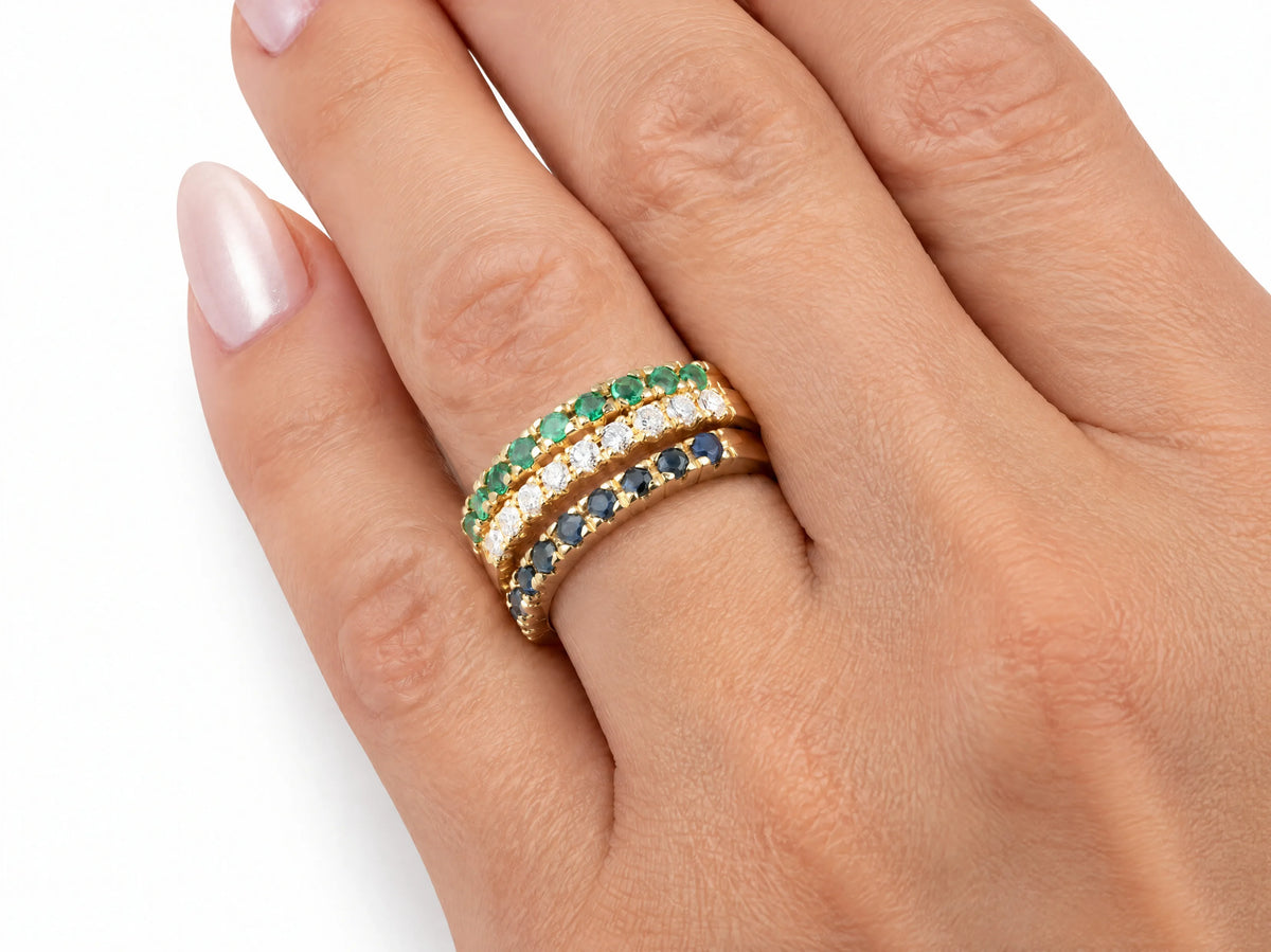 ArtGold 18K yellow gold half eternity bands worn together on the finger — diamond, emerald and sapphire, each set with nine round stones