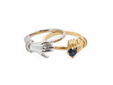 Angled view of two-tone gold ring with separated sculpted hands reaching toward sapphire