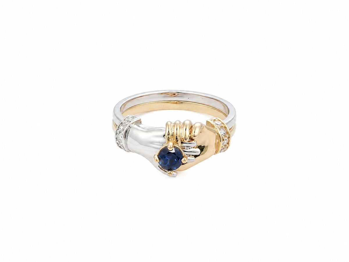 Front view of two-tone gold ring with two sculpted hands united, cradling sapphire and diamond accents