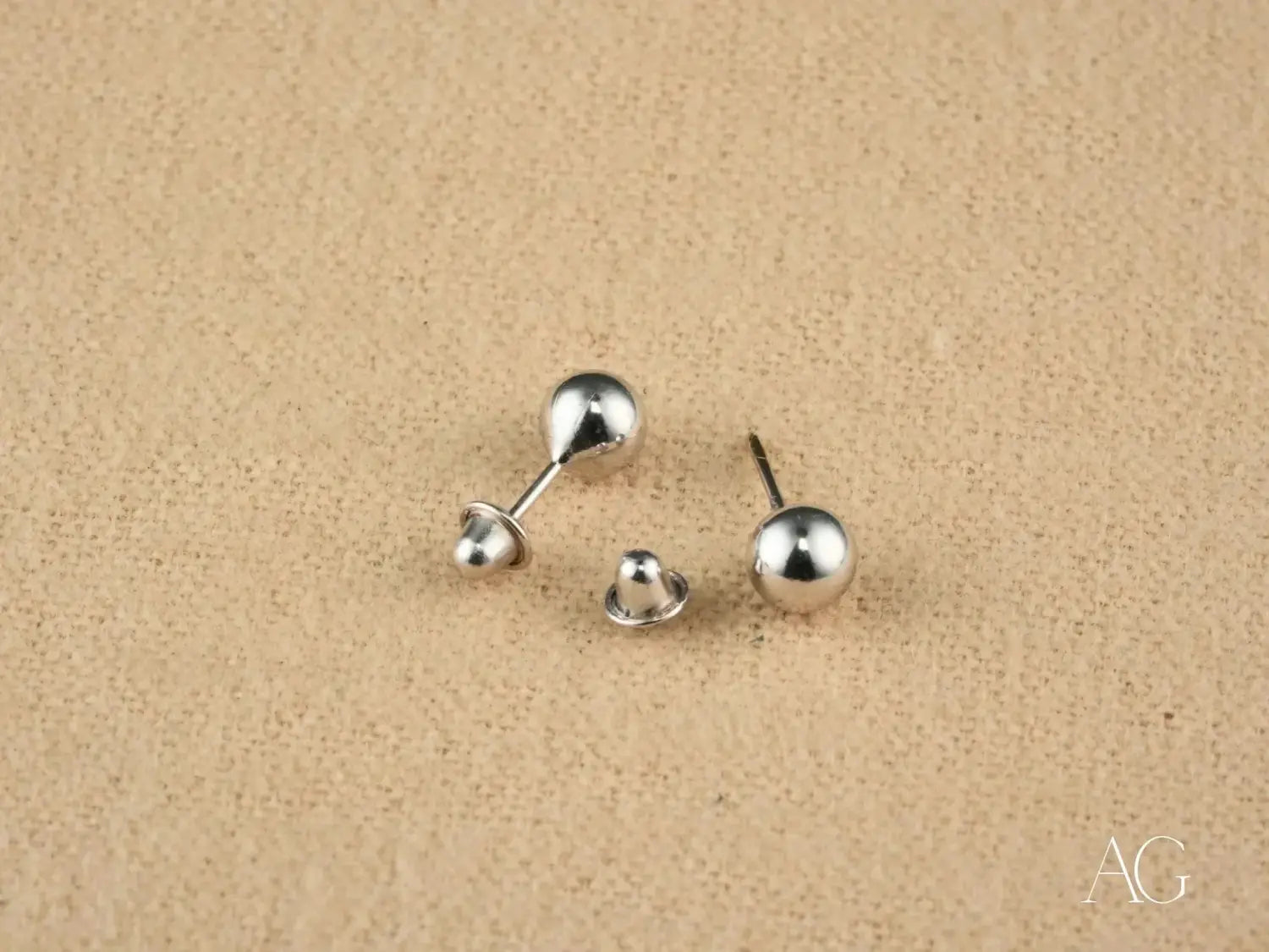 a pair of sleek minimalist silver ball stud earrings with a polished metallic finish featured in an article comparing buccellati and de beers in the evolution of fine jewelry focusing on gold jewelry and 18k gold craftsmanship