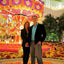 Two people posing in front of a colorful, floral carousel and flower display.