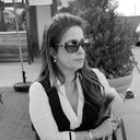 A woman wearing sunglasses, a black vest over a white shirt, and multiple pieces of jewelry.